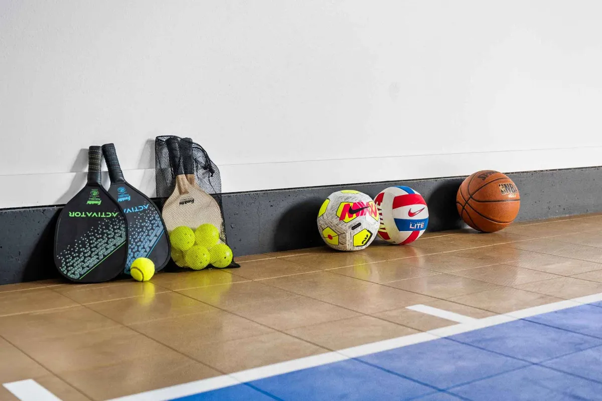 A vibrant indoor sport court scene featuring paddles, tennis balls, soccer balls, volleyball, and basketball, with ample space for a variety of active games. The setting exudes energy and is clearly arranged for family-friendly play, regardless of weather.