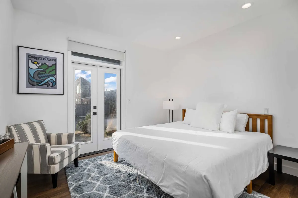 A tranquil bedroom with a bed, elegant artwork, sliding glass doors that open to natural light, and a comfortable armchair. The space feels peaceful and offers a sense of private sanctuary, perfect for recharging after a day at the coast.