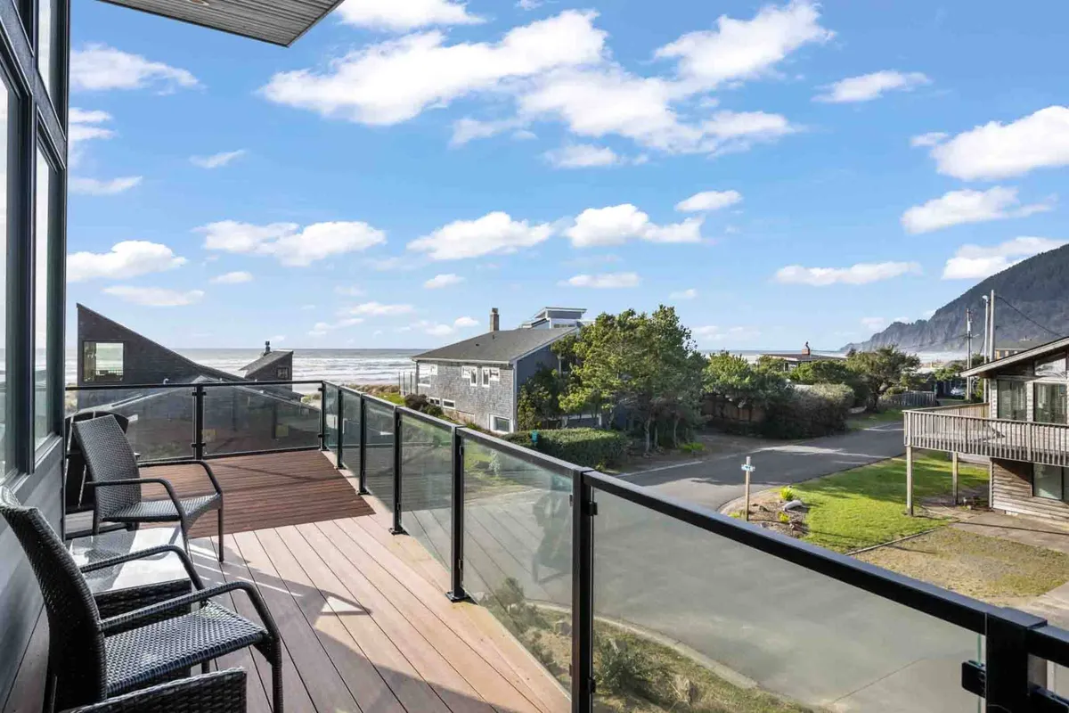 A tranquil balcony with a railing overlooks expansive ocean views, with the blue horizon stretching beyond surrounding homes and a wide open sky. The setting is perfect for relaxing, with the promise of both sunrise and sunset vistas.