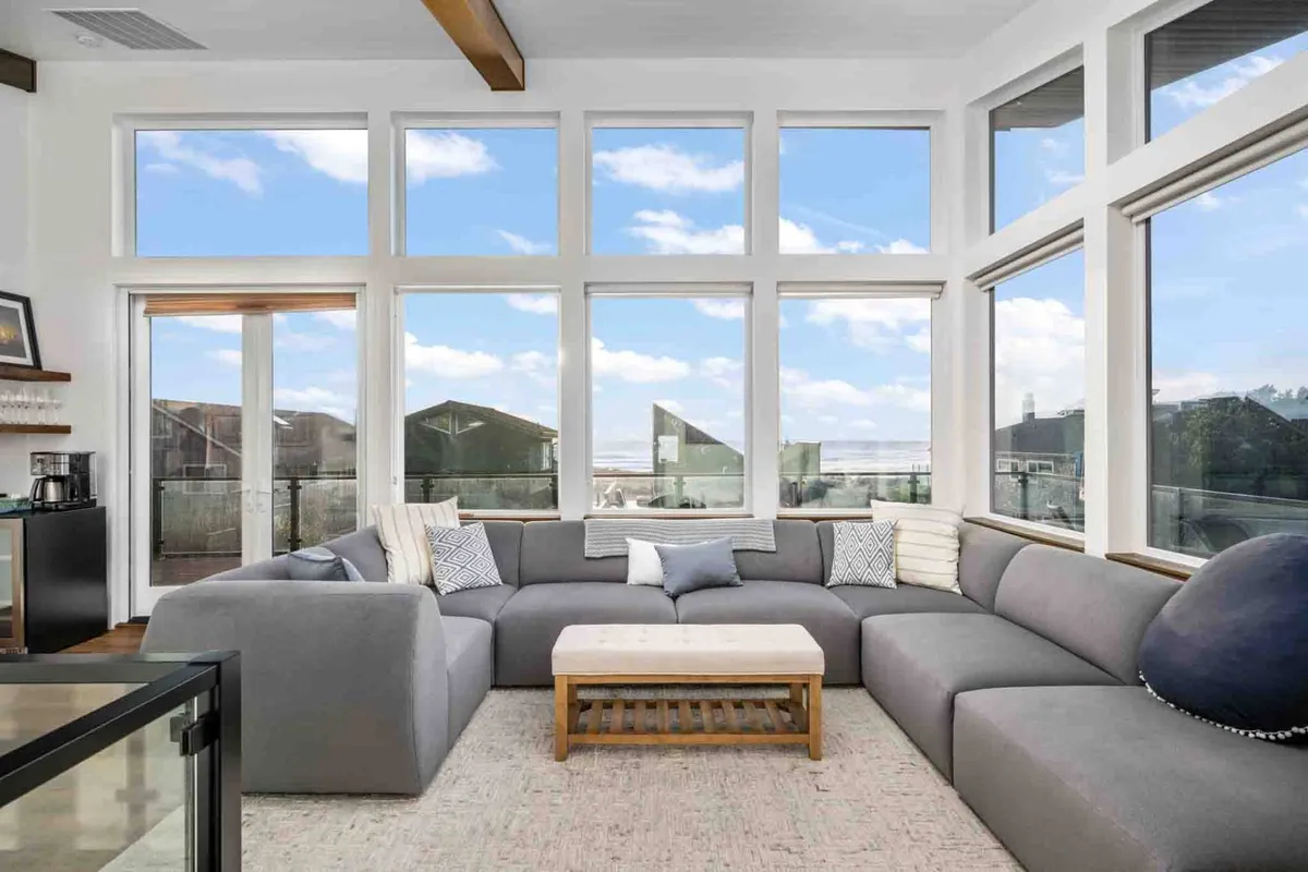 This image showcases a spacious, open-plan living room featuring a large sectional sofa and a coffee table, all positioned to face expansive floor-to-ceiling windows. The windows flood the space with natural light and offer scenic views of the Pacific and Neahkahnie Mountain. The room is arranged for relaxing, socializing, and enjoying the sunset.