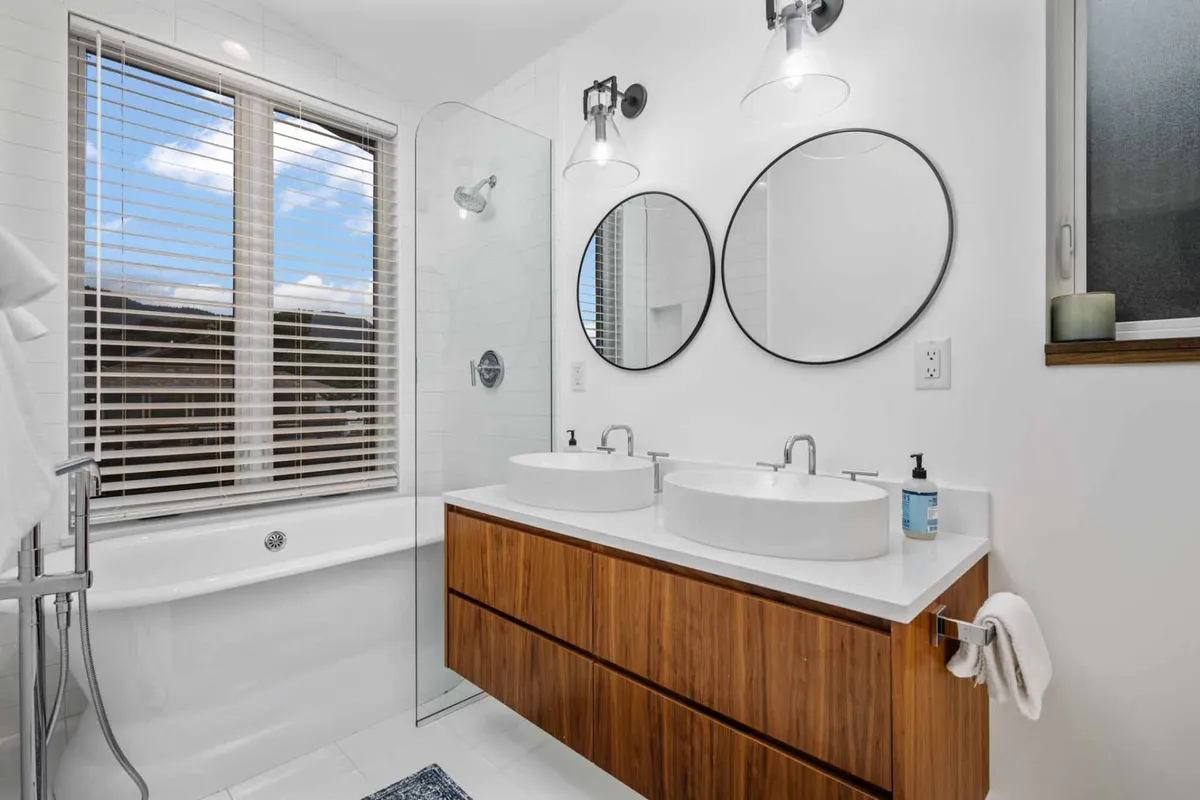 A modern bathroom with double sinks, large mirrors, a bathtub, and a window with blinds. The space is organized, airy, and filled with natural light, evoking a spa-like mood that invites relaxation and self-care.