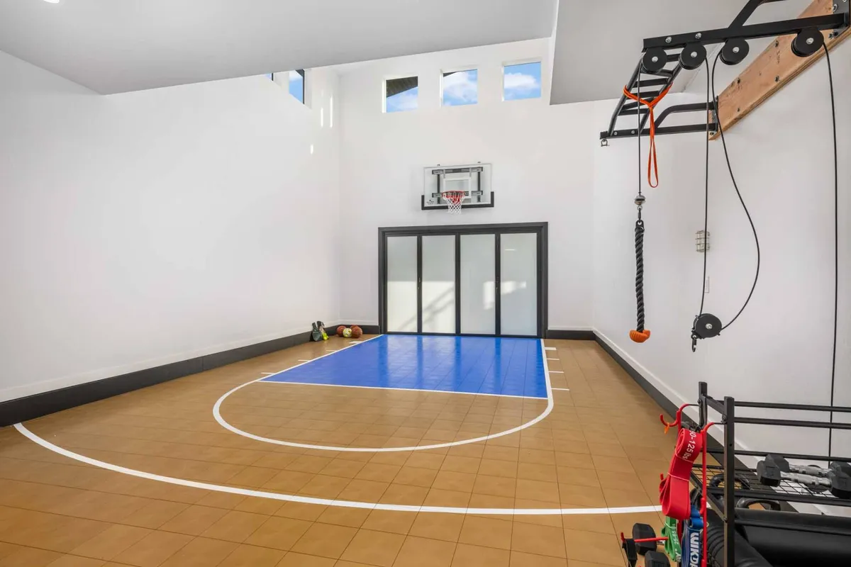 A lively interior shot showcasing the indoor half-sport court with a basketball hoop, exercise equipment neatly arranged along the wall, and sunlight streaming through large windows. A family is enjoying a basketball practice session, while others are using the workout equipment, highlighting the space's versatility for both play and fitness.
