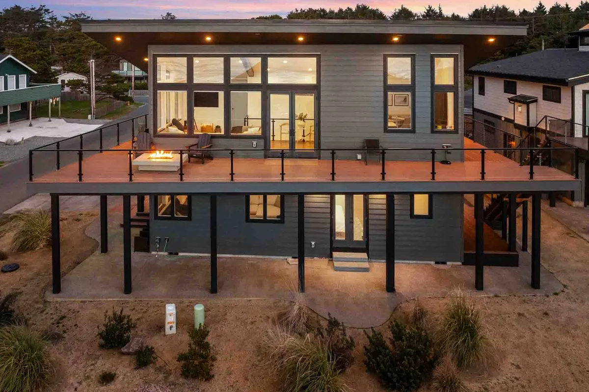 An inviting outdoor deck highlights large windows, stylish outdoor seating, and a fire pit. The view extends to the horizon, with soft evening light and a hint of coastal breezes.