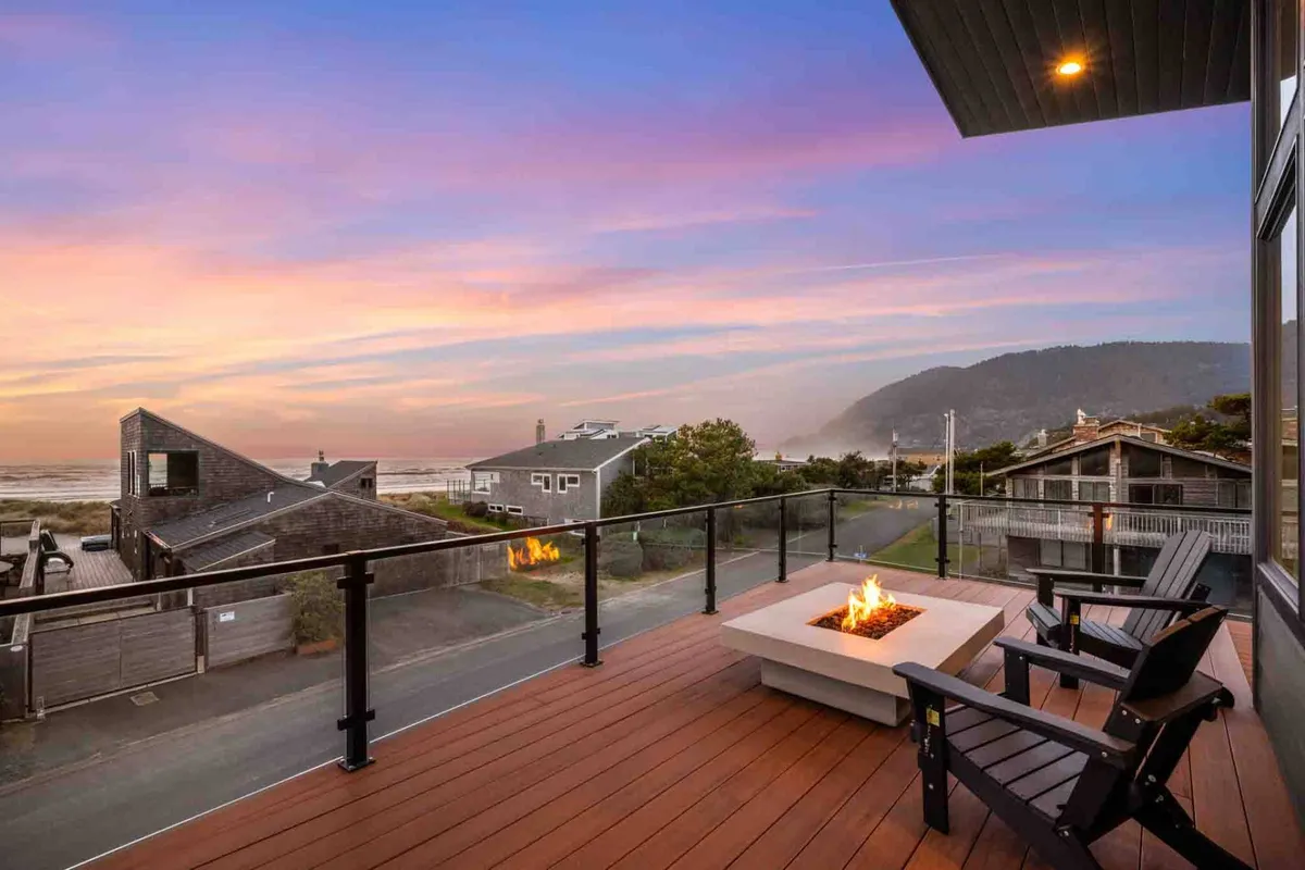 The image showcases a stylish outdoor deck overlooking the ocean at sunset, with comfortable chairs arranged around a glowing fire pit. The serene sky is painted in warm hues, inviting relaxation and connection as guests gather to enjoy the evening view.