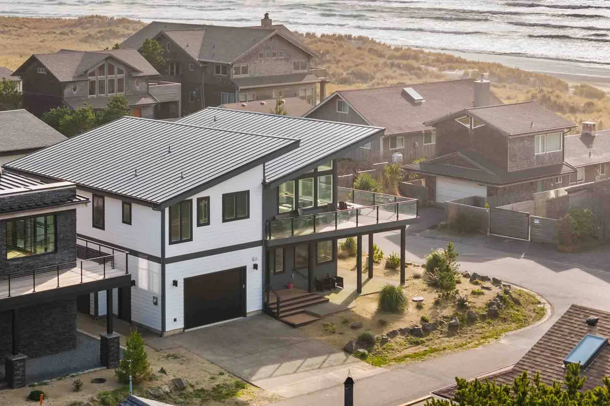A wide-angle exterior shot features the home’s modern architecture with large windows, an ample deck area furnished for gatherings, and a landscaped yard. The space is clearly designed for family and friends to enjoy barbecues and coastal scenery.