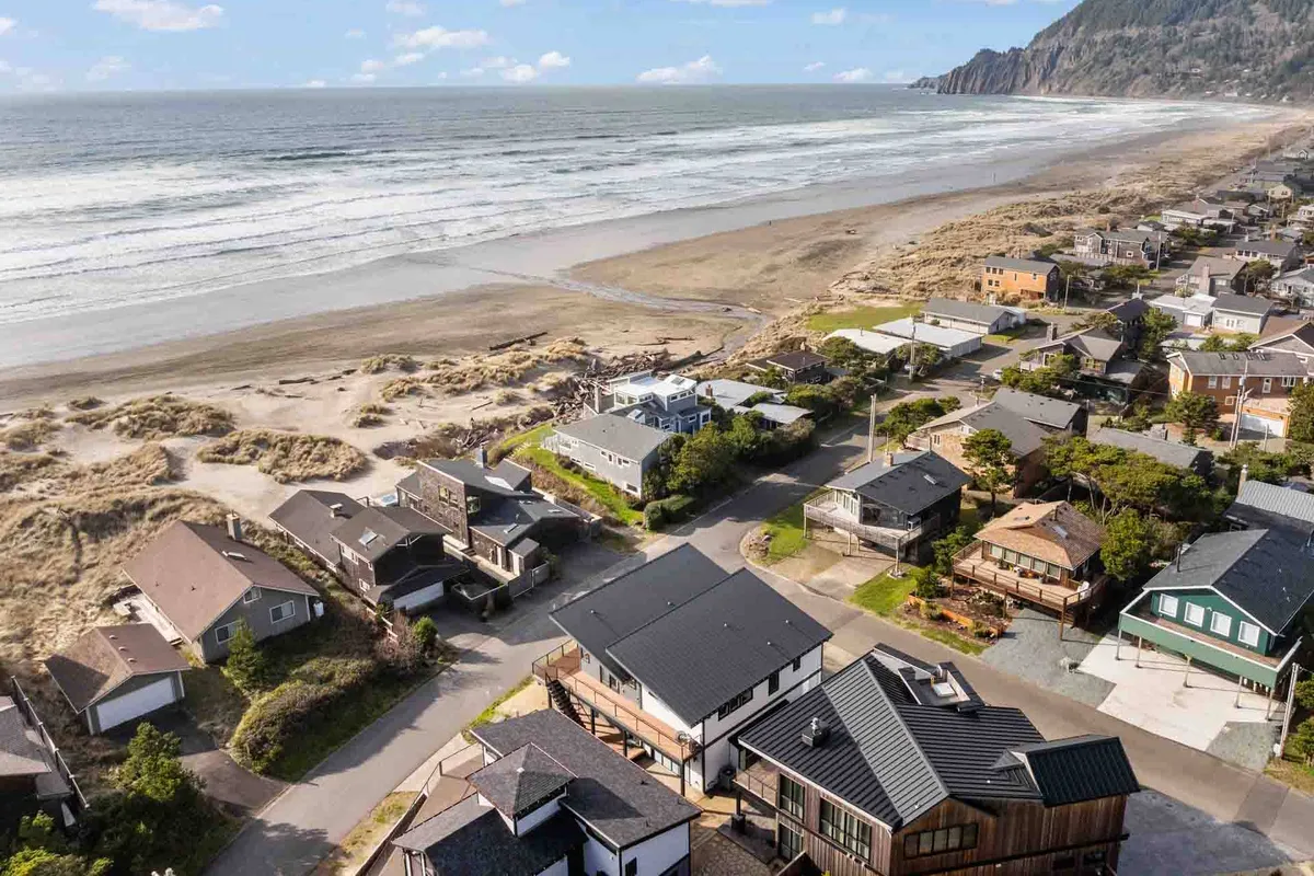 A sweeping stretch of Manzanita Beach is captured with rolling ocean waves, golden sand dunes, and beachfront houses nestled below cliffs in the background. The morning light highlights the wild, natural beauty of the shoreline, inviting exploration and relaxation.