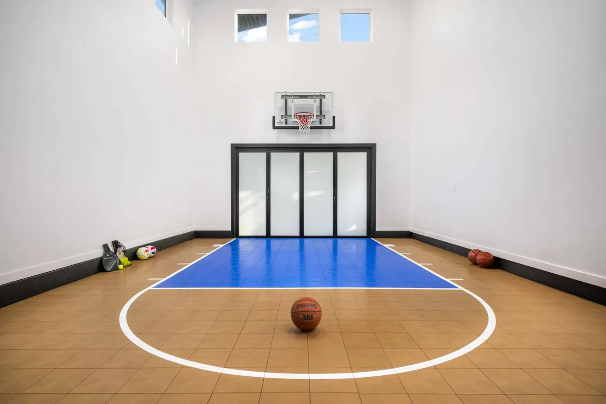 A playful close-up of the basketball hoop, basketball, and other sports balls like soccer and tennis, scattered across the polished indoor court. The scene suggests ongoing games, celebrations, and moments of learning and fun for all ages.