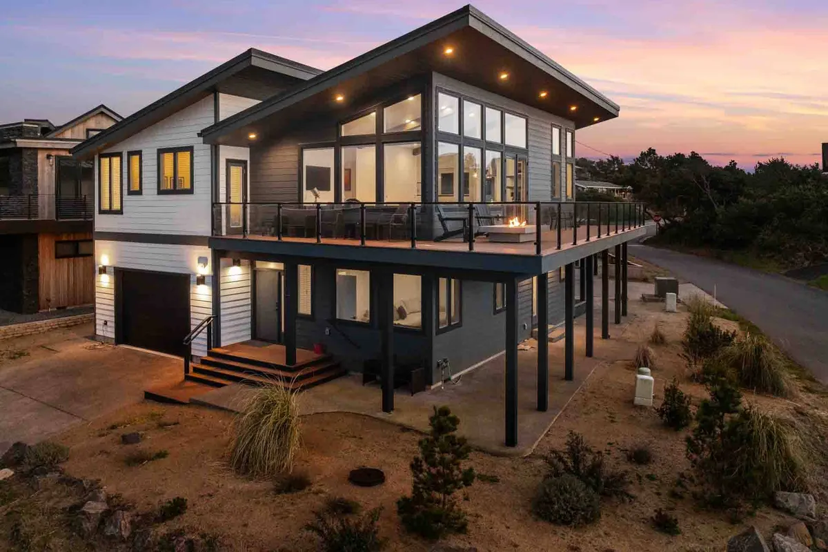 This exterior image features a modern deck with outdoor seating, set against large glass windows. The scene is tranquil and inviting, with the deck designed for gatherings, relaxation, and sharing stories around a fire pit as the sun sets over the horizon.