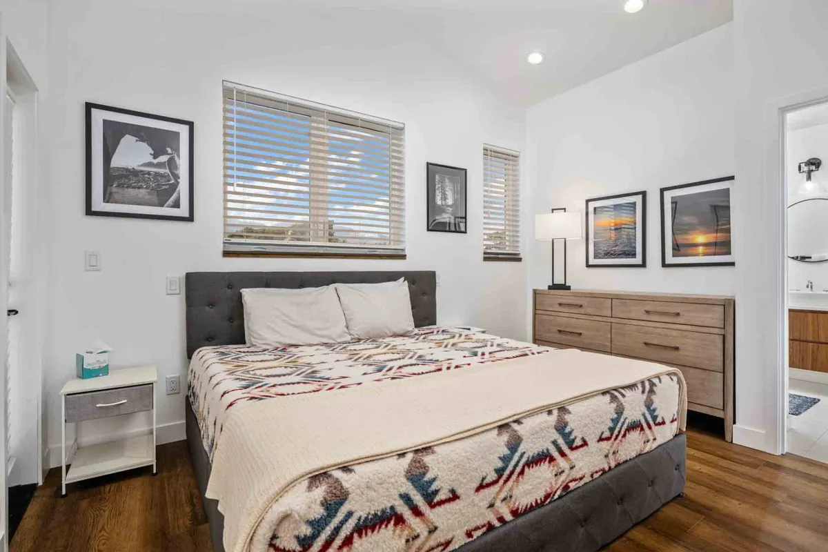 A cozy bedroom with a bed featuring a patterned blanket, framed pictures on the wall, and a nightstand with a lamp. Natural light streams in through a window, creating a serene and inviting atmosphere ideal for relaxation.