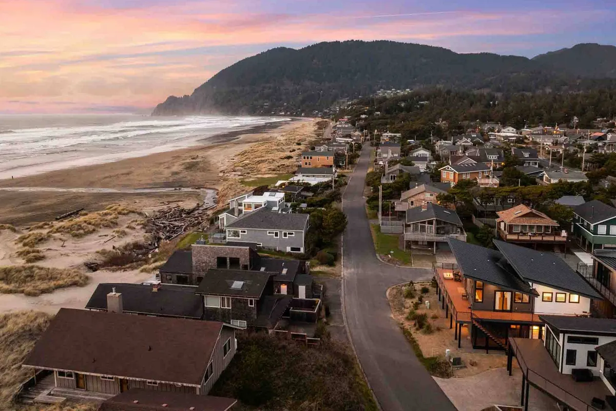 A sweeping coastal view of the beach, ocean, nearby homes, and mountains in the distance. The scene captures expansive sandy shores ideal for walks, sandcastle building, and kite flying, with the natural beauty of the coast inviting outdoor exploration.