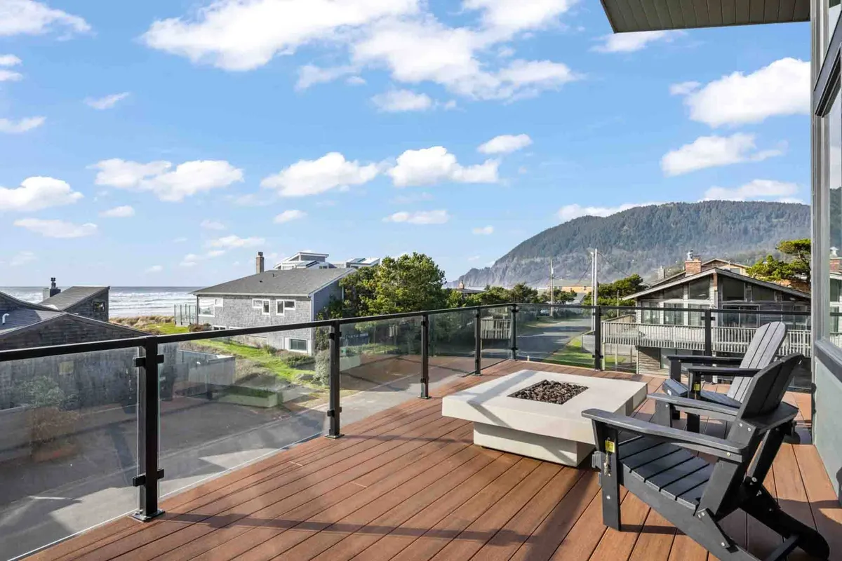 A tranquil deck scene with comfortable deck chairs circling a modern fire pit, all positioned to take in expansive ocean views against a mountain backdrop. The golden sunset light bathes everything, suggesting both relaxation and socialization.