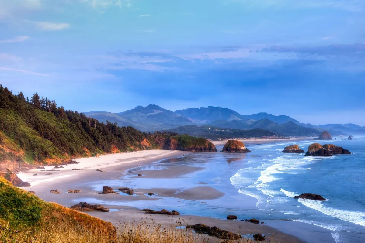 A breathtaking natural scene featuring waves crashing against rugged rock formations, with distant mountains rising along the horizon. The shoreline is wild and inviting, suggesting ample opportunities for beachcombing, hiking, and photography.